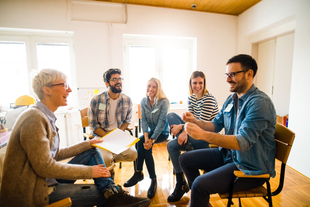 A man speaks in a circle of people, who are all laughing.