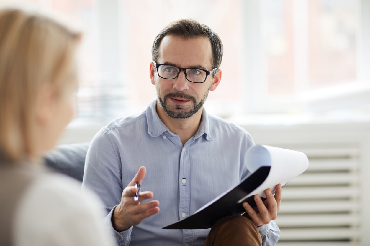 A man with clipboard and pen gestures and talks to a woman sitting opposite him.