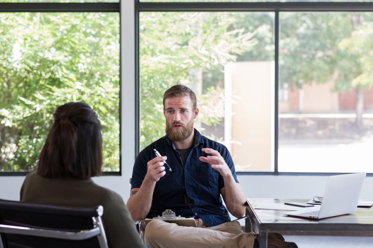 A serious therapist with clipboard gestures and talks to woman sitting opposite him.