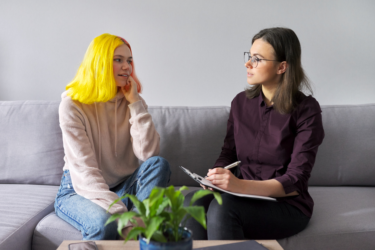 A teenage girl talks to a therapist with a notepad on sofa.