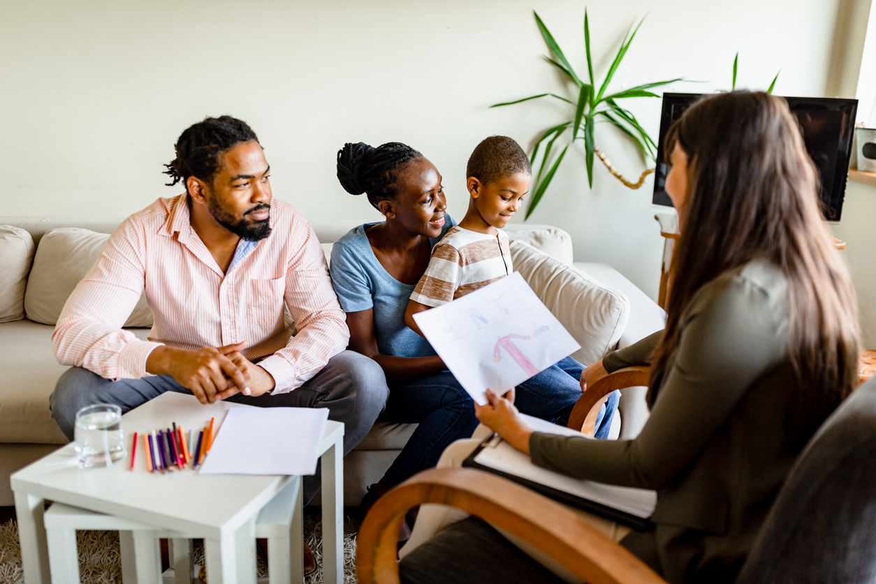 A couple sit looking at their smiling young son on a sofa while a therapist examines his drawing.