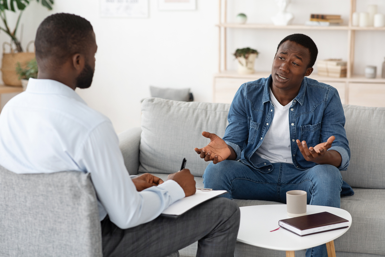 An anxious man gestures as he talks to a therapist across from him who takes notes.