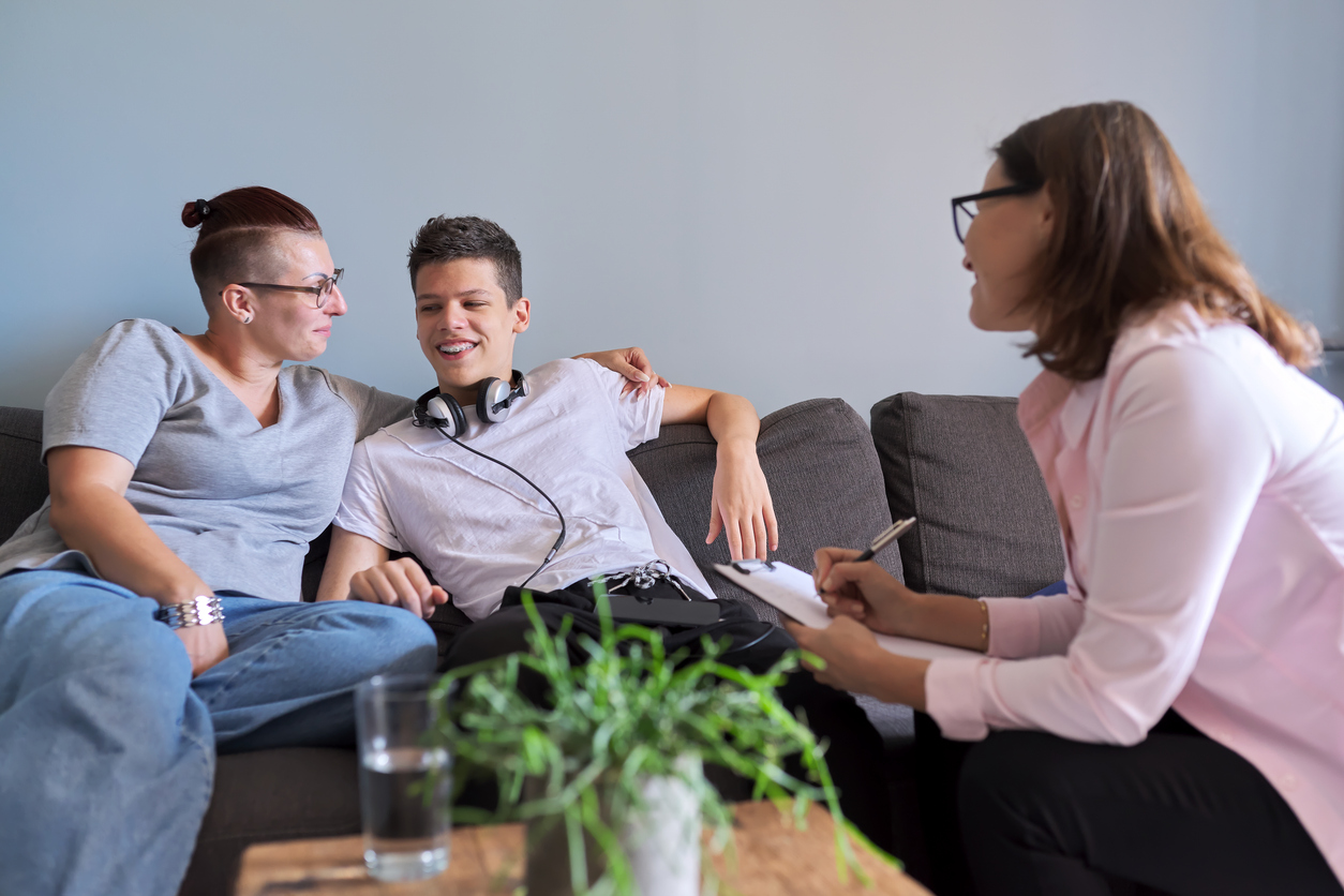 A mother and teenage son sit on a sofa talking a woman, who smiles and makes notes.