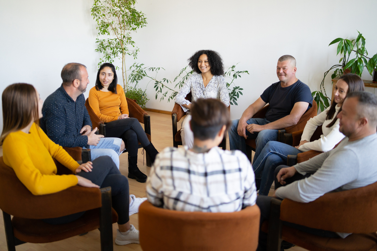 A circle of men and women sit in comfortable chairs talking and smiling.
