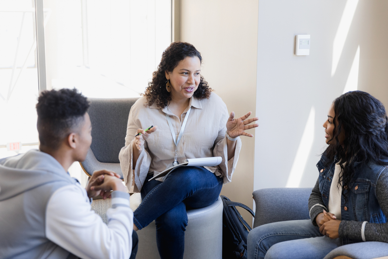 A therapist with a notepad talks to a young man and young woman in an office.