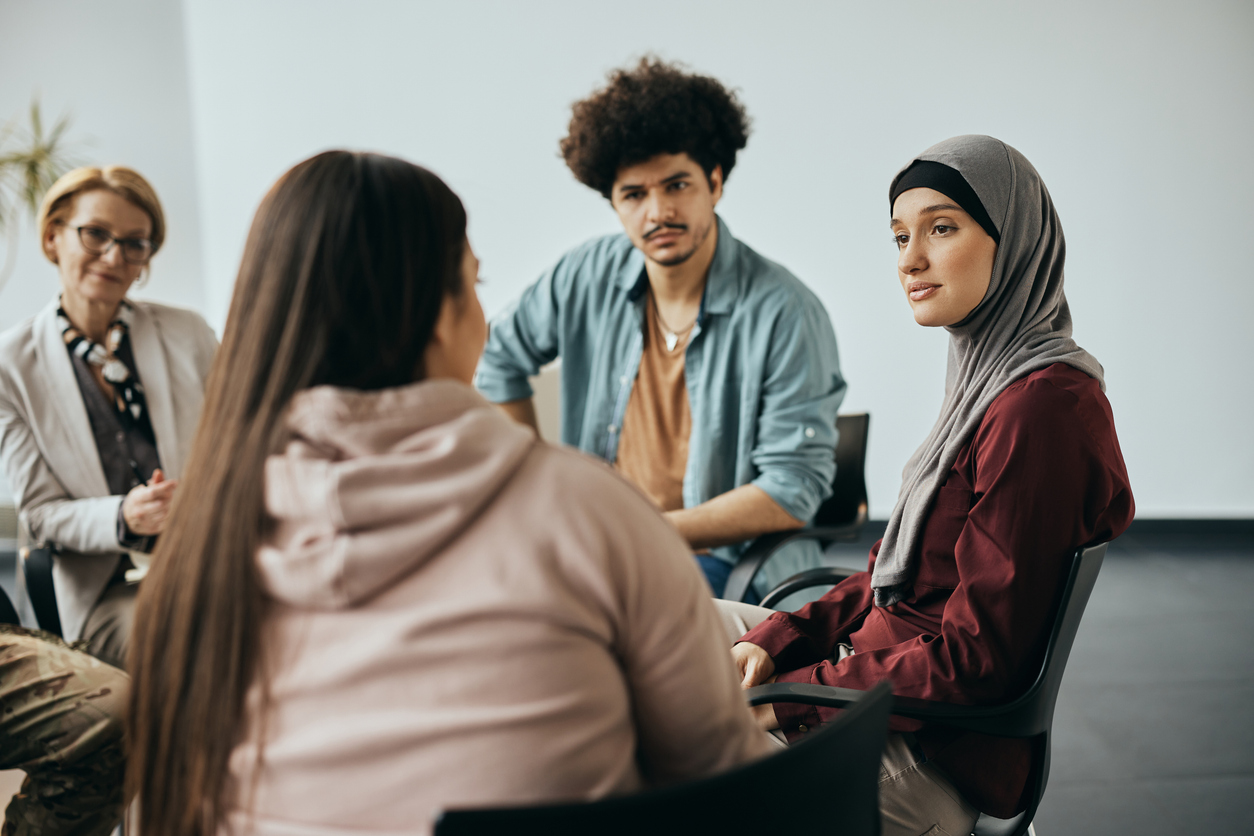 A circle of people in a group therapy session listen attentively as one of them speaks.