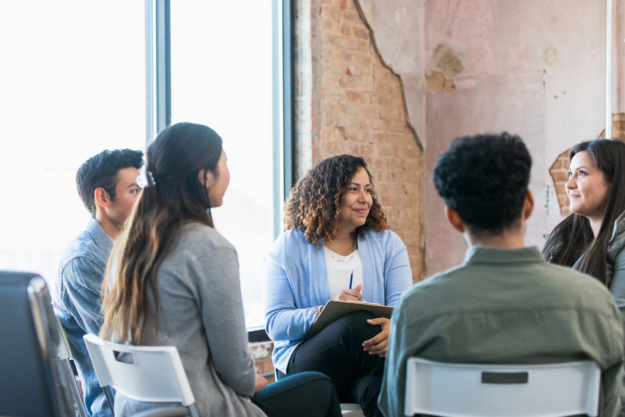 A small circle of men and women sit listening to a young woman while a therapist prepares to make notes.