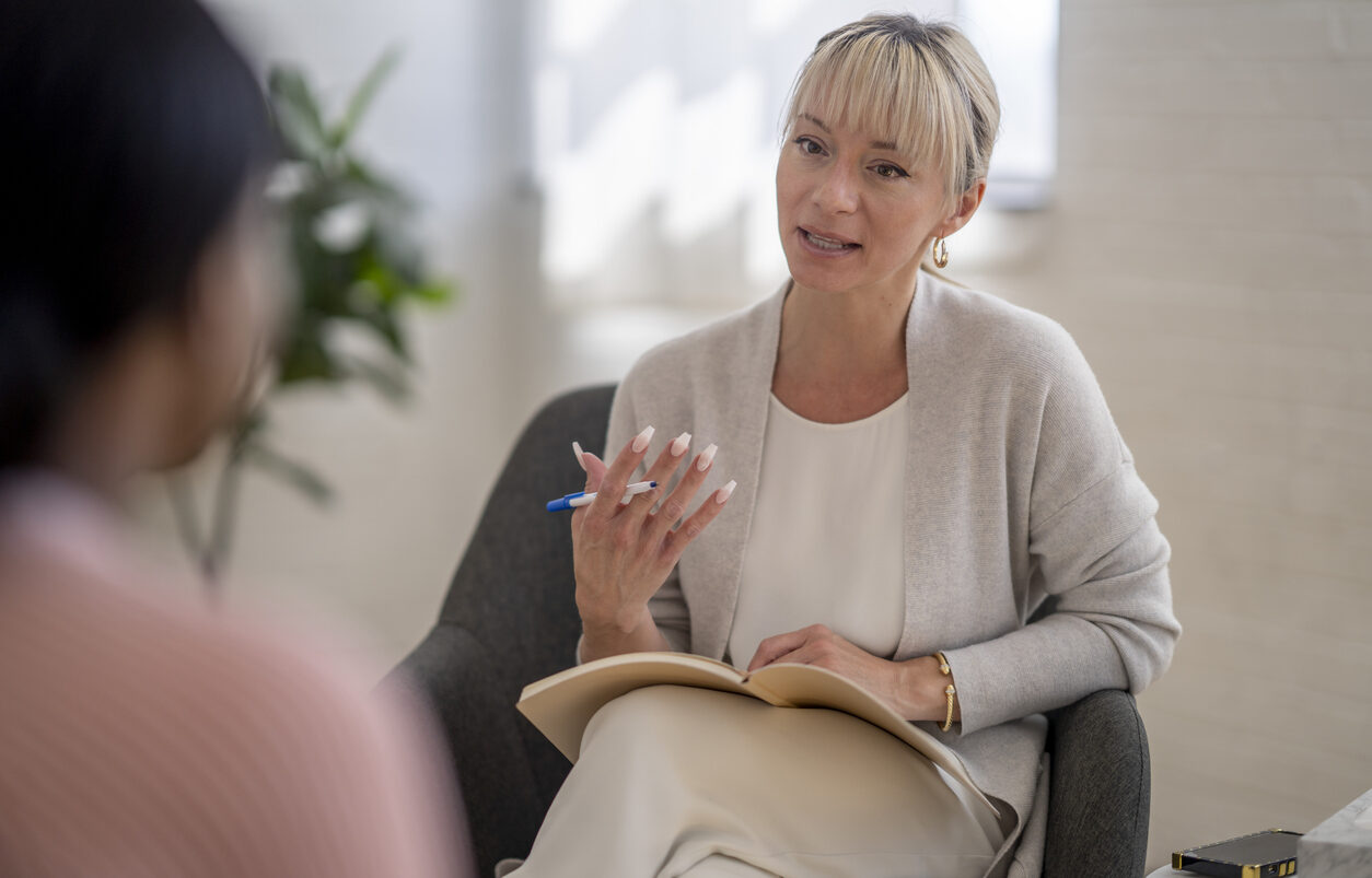A therapist with a notebook gestures and talks to the woman sitting opposite her.