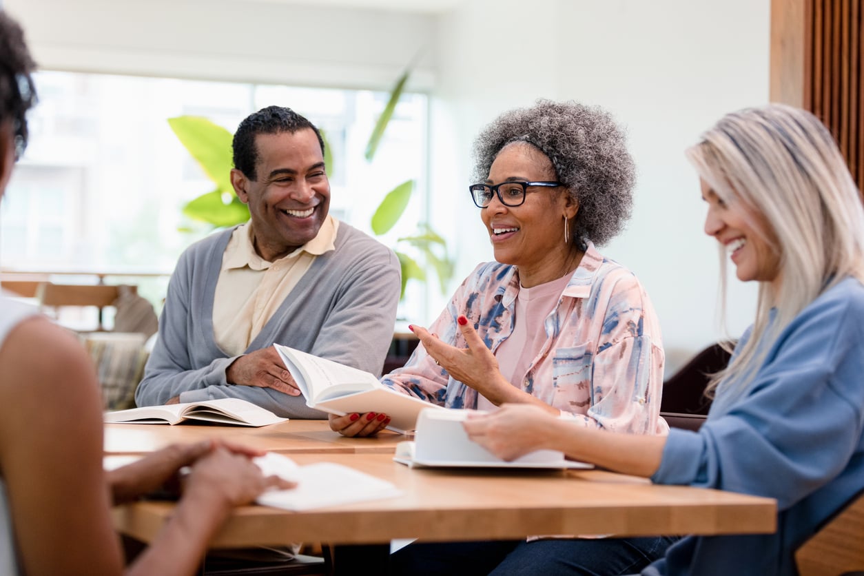 A woman at a table talks to smiling people with books in front of them.