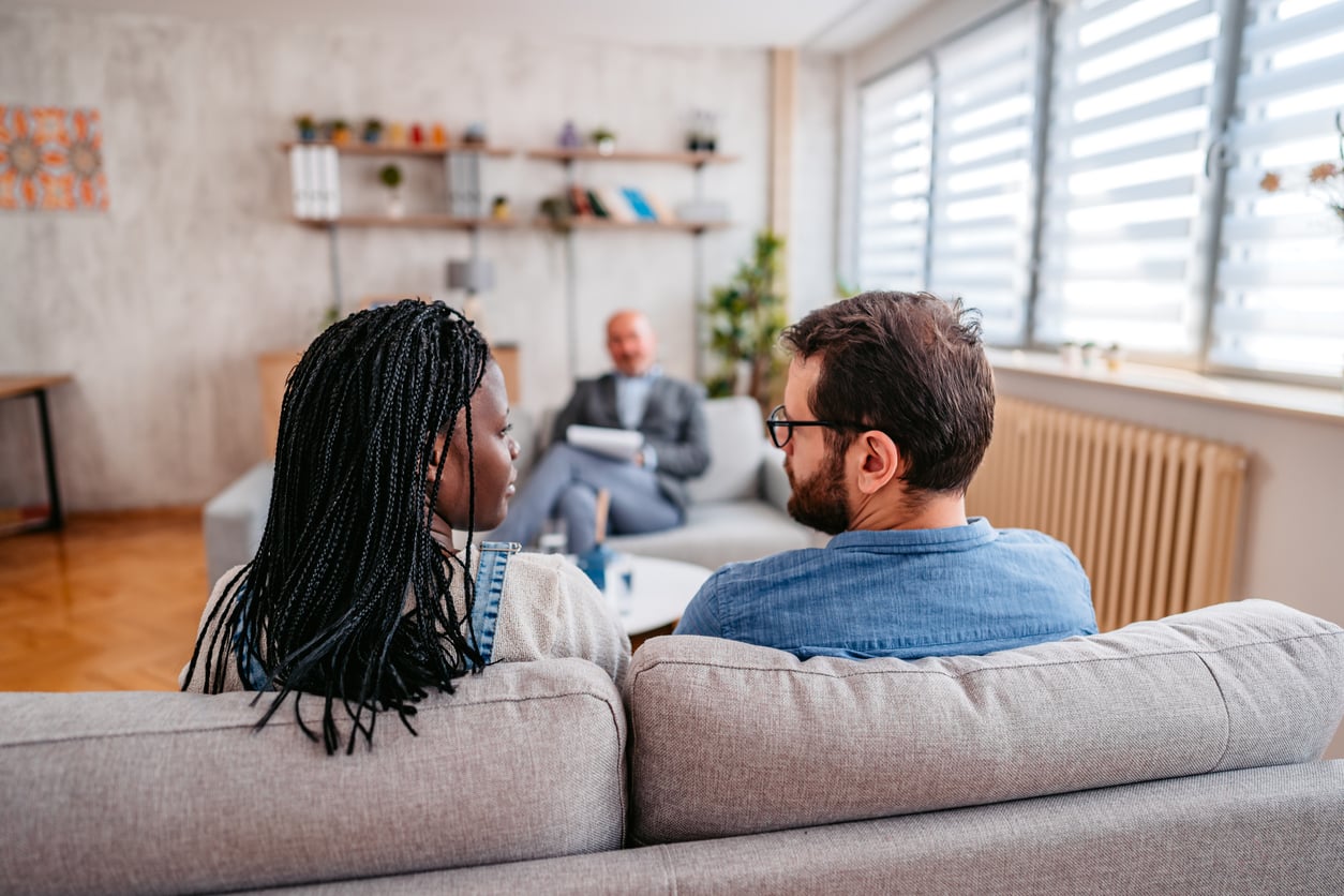 A couple sitting on a sofa look at each other while a therapist opposite them makes notes.