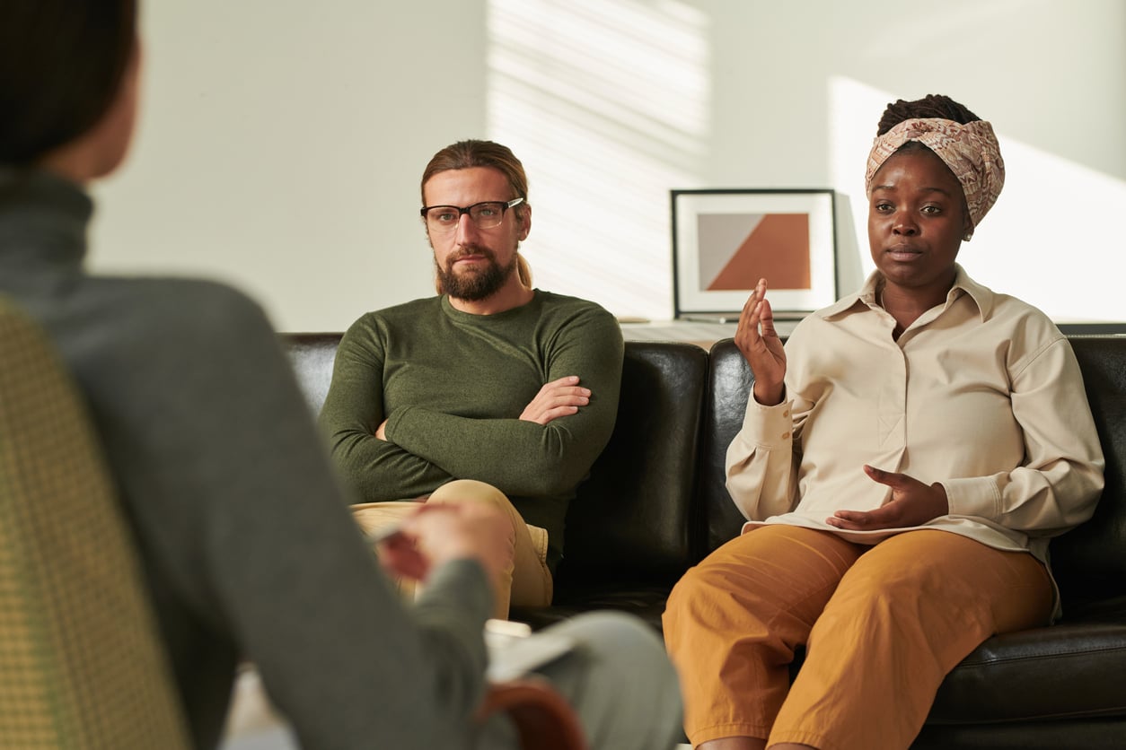 A couple sit on a sofa talking to a therapist.