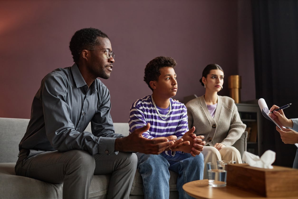 Two parents and their teenage son sit on a couch, talking to a therapist who is taking notes.
