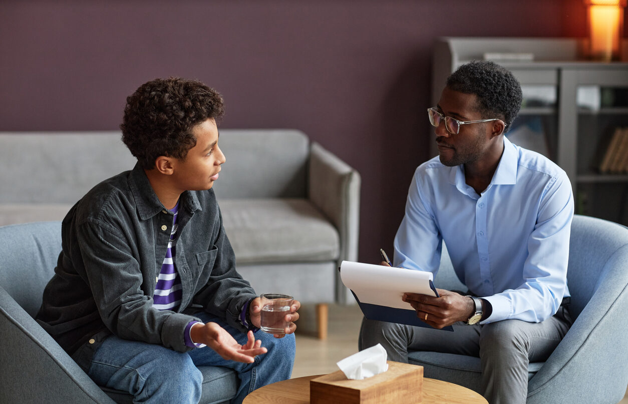 A therapist makes notes as he listens to a teenage boy, who is holding a glass of water.