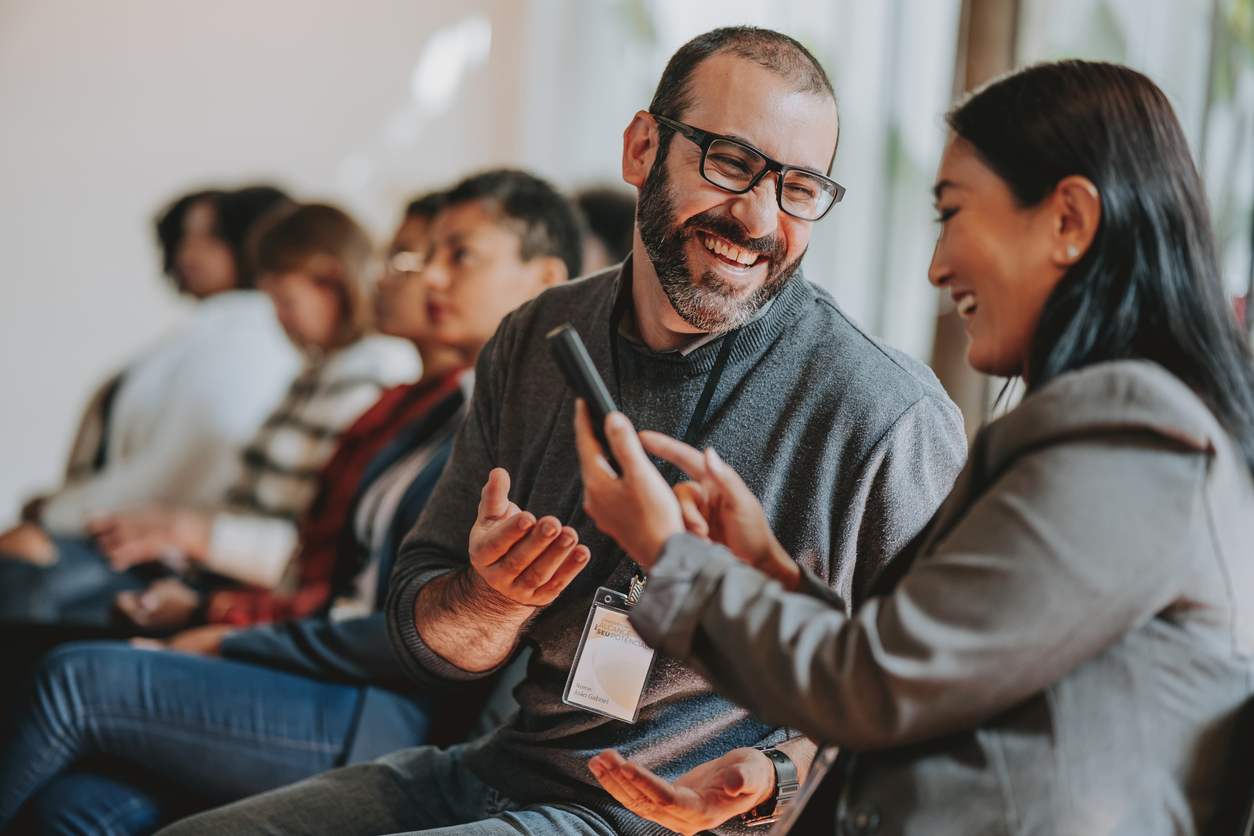 A woman shows a man something on her smartphone at an event, and they both laugh.