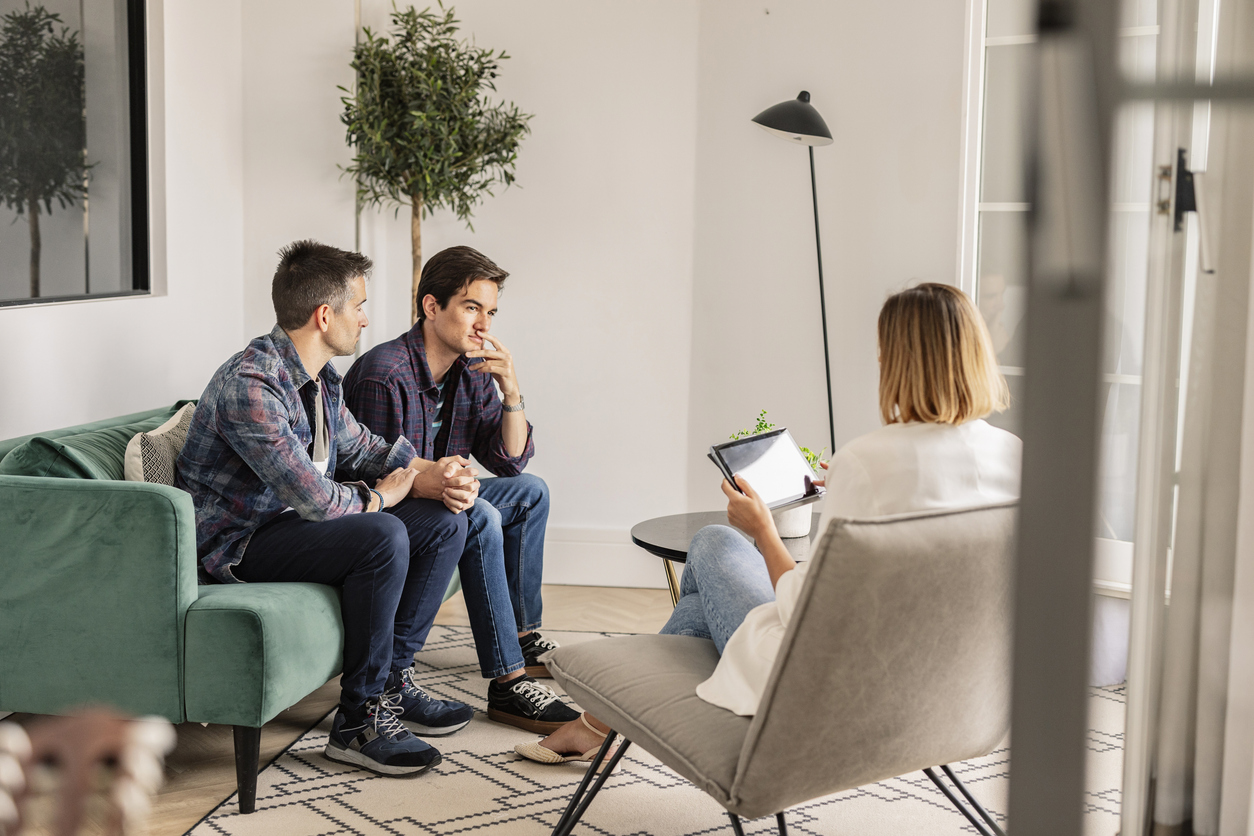 A serious looking gay couple sit talking to a therapist, who is looking at notes on tablet.