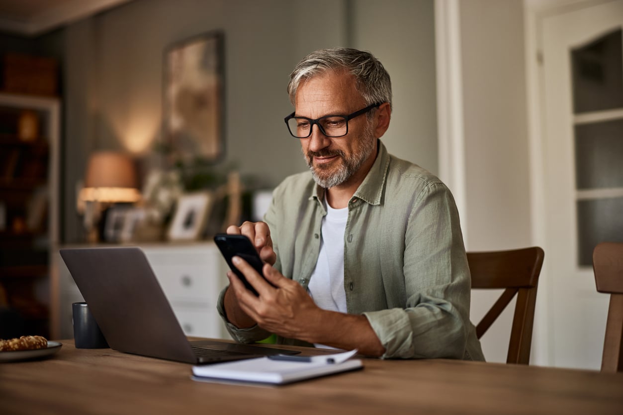 A man at a table with a laptop in front of him checks something on a mobile phone.