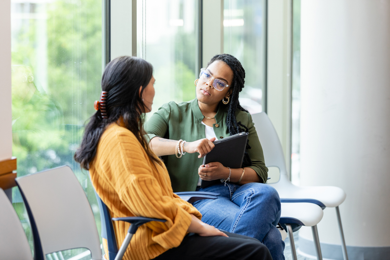 A therapist holding a laptop listens to a woman sitting next to her.
