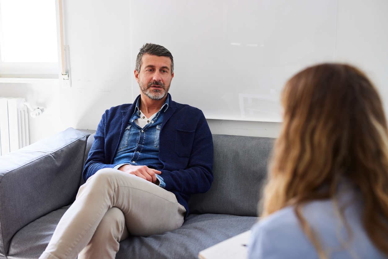 A serious looking man sits on a sofa talking to a therapist, who is making notes.