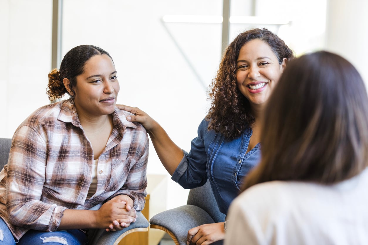 A woman smiles and places her hand on another woman's shoulder while they talk to a therapist sitting opposite them.