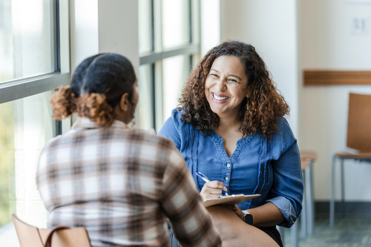 A young woman talks to a therapist, who is making notes and smiling.