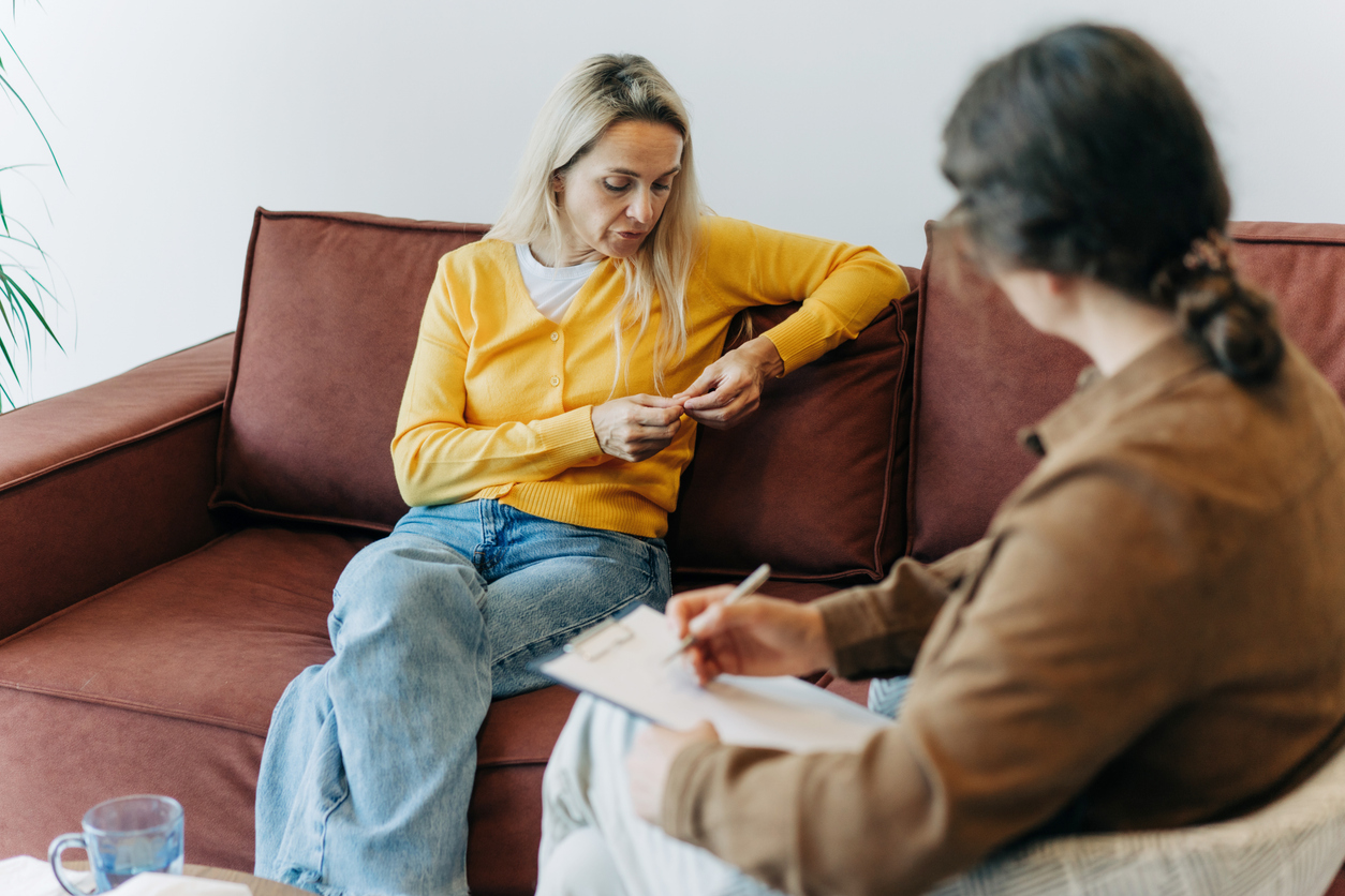 An unhappy looking woman sitting on a couch looks at her hands while talking to a therapist, who is making notes.