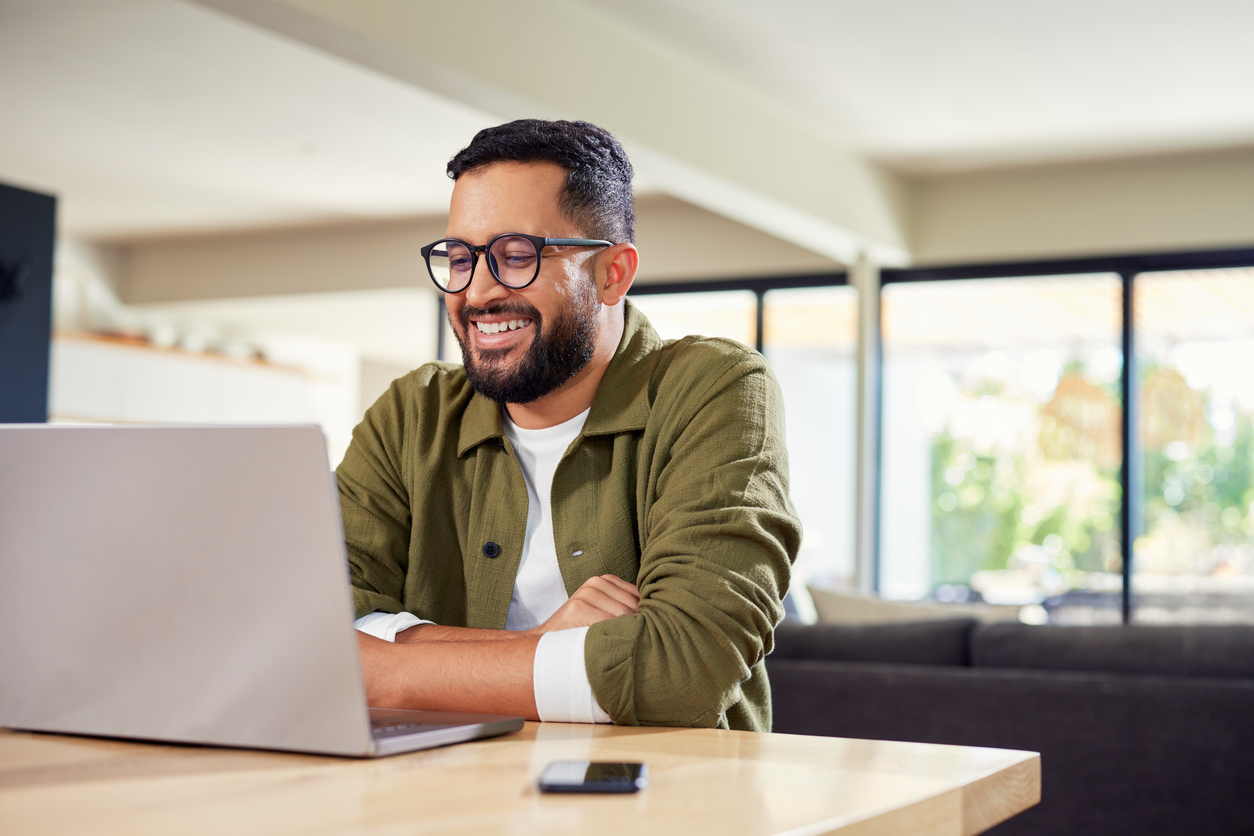 A man sitting at a table looks at a laptop and smiles.